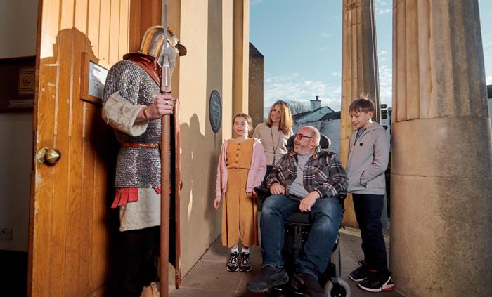 A man in gladiator costume stands to the left of the picture. He is meeting a family at the front entrance of the museum. It is a grand entrance with